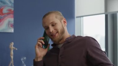 Rahatlamış bir adam ofis portresinde akıllı telefonlardan bahsediyor. Gülümseyen serbest çalışan, mavi duvar odasındaki dinlenme koltuğunda telefonla konuşuyor. Modern uzman, penceredeki iş yerinde mola veren arkadaşlarıyla konuşuyor