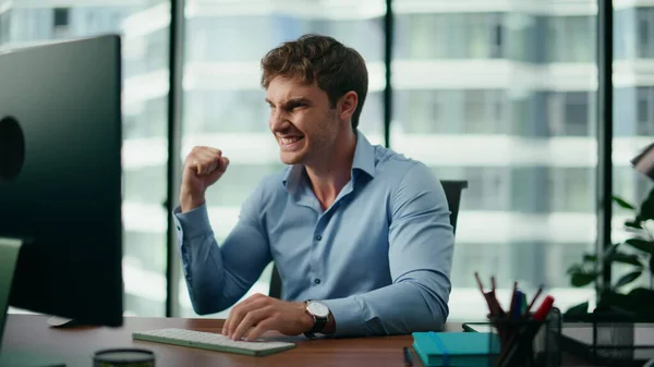 Happy businessman celebrating triumph emotionally sitting at office table modern skyscraper close up. Portrait of excited worker reading on computer screen positive news. Man enjoy career achievement.