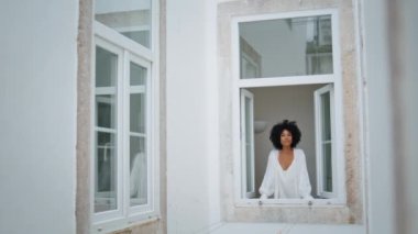 Pozitif kadın pencereye bakıyor. Kıvırcık gülümseyen kadın dışarıda dikizlemekten hoşlanıyor. Mutlu Afro-Amerikan modelleri sabah şehir manzarasının rahatlamasını seyrediyor. Günlük rutin kavram