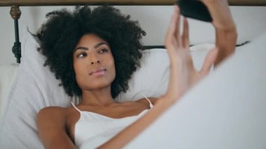 Yatakta Afrikalı bir kadın selfie için poz veriyor. Beyaz yastıkla kendi portresini yapan yakın plan neşeli kız. Gülümseyen kadın otel yatağında dinlenirken fotoğraf çekiyor. Modern kadın yaşam tarzı konsepti