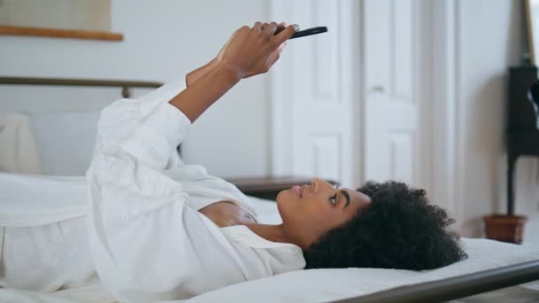 把女人的留言机放在卧室近旁 卷发黑发发女郎在豪华别墅的侧视图中发短信休息床上 一个拿着智能手机独自浏览社交媒体的非洲裔美国女孩 — 图库视频影像