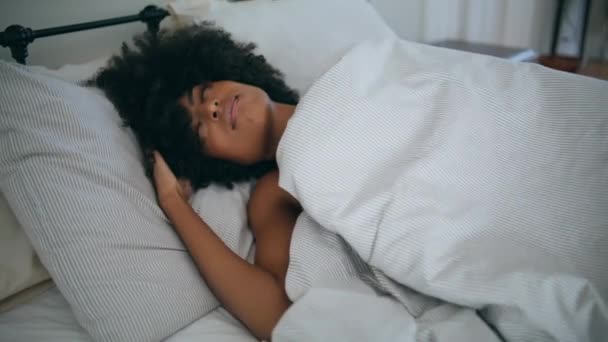ベッドで目が覚めるモデル 毛布の下に早朝に隠れて枕を敷くカーリーのヘアスタイルの眠い女性 アフリカの深刻な女の子は週末に柔らかいマットレスで休んでいます 女性怠惰なライフスタイルの概念 — ストック動画