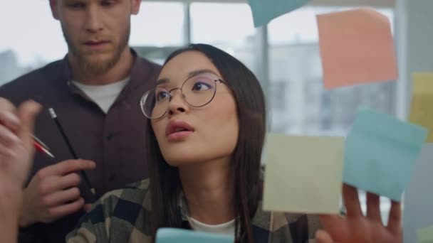 オフィスで眼鏡作成者の書き込みステッカーの壁を閉じます アジアの女性は一緒にアイデアを共有する男の同僚と話している ホームでのブレインストーミングセッションでは 付箋を使ったスタートアップチームが参加 — ストック動画