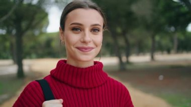 Yeşil ormanın önünde duran, kırmızı süveter giyen sevimli genç bir kadının portresi. Çekici, mutlu, çantalı bir kız doğada yürüyecek. Yakından. Güzel, esmer, gülümseyen kamera..