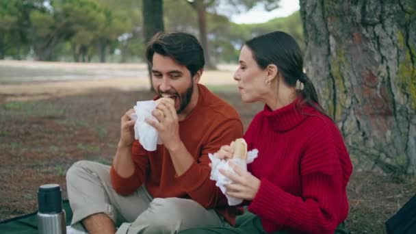 秋の公園でピクニックでサンドイッチを食べる若いロマンチックな家族が閉じます 明るく幸せなカップルは屋外の毛布の上に座っておいしい軽食をお楽しみください 森の中で笑顔の男とリラックス魅力的な女性 — ストック動画