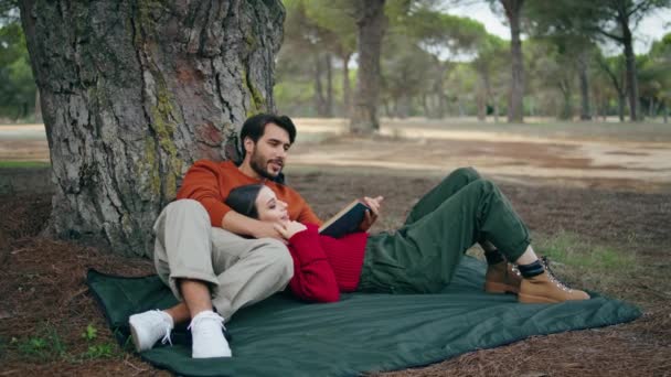 ロマンチックな森のピクニックで毛布の上に寝そべって公開カップル 魅力的なひげを生やした男が大声で本を木にもたれて読んでいる ボーイフレンドの胸を屋外でリラックスして介護無料の若い女性 ファミリーレジャーのコンセプト — ストック動画