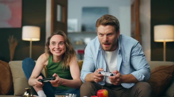 活跃的朋友在舒适的房子里玩着游戏机 兴奋的夫妇牵着控制器试图在视频游戏中获胜 专心致志的男人微笑的女人享受比赛 在沙发上消磨时光 — 图库视频影像