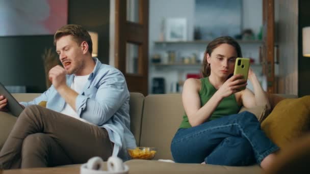 ソファで保持しているインターネット中毒ペアガジェット 本格的な女性はソファでリラックス電話を見て 集中的な男は自宅でタブレットPcの電子書籍を読んでチップを食べる 現代の家族の週末のコンセプト — ストック動画