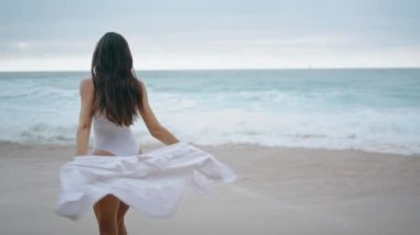 Kaygısız güzel kadın kumlu sahilde beyaz seksi bir mayo giyerek dönüyor. Köpüklü okyanus dalgalarının önünde yürüyen çekici, ince kız. Mutlu bayan bulutlu deniz kıyısında yaz tatilinin tadını çıkar..