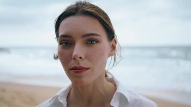 Güzel, nazik kadın yüzü kameraya dikkatlice bakıyor. Deniz dalgalarına yakından bakıyor. Bulutlu sahilde tek başına beyaz gömlek giyen çekici ve düşünceli bir kızın portresi. Deniz kıyısında poz veren sakin bir bayan.