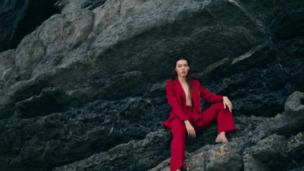 赤い挑発的なスーツを着て山の棚に座って美しい女性を自信を持って 石の岩の屋外の曇りの週末にポーズ豪華なセクシーなモデル 本気な女の子見ますカメラ上の自然 — ストック動画