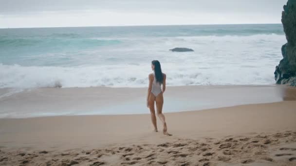 性感的小女孩穿着白色泳衣站在沙滩上 一个不知名的迷人女人走在充满泡沫的海浪的阴天 无忧无虑的女士享受美丽的海景阴郁的一天 — 图库视频影像