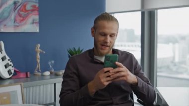 Ofiste akıllı telefonlara mesaj atan rahat bir adam. Gülümseyen serbest çalışan koltukta dinleniyor mavi duvar odasında internete takılıyor. Modern iş adamı vitrinlerde sosyal medyayı izlerken mola verdi