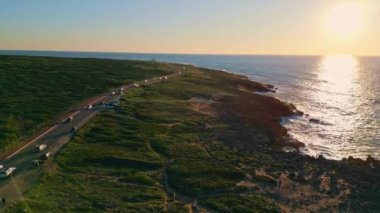 Yazın gün batımında, kayalık sahillerde sahil yolu. Deniz kıyısında ağır çekimde giden arabaların sinematik drone görüntüsü. İnanılmaz gün batımı dalgalı okyanus yüzeyine yansıyor. Turizm konsepti.