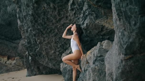 石の岩にもたれて海岸にポーズをセクシーな若い女性 海岸に白い水着姿の魅力的な官能モデル 崖の近くのビーチの砂の上に立つ熱い豪華な女の子 美の概念 — ストック動画