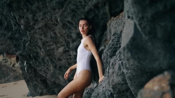 熱い若い女性岩の海岸の石の崖にもたれてポーズ 白いセクシーな水着を自信を持って着てカメラを見て魅力的な官能的なモデル 美しい真剣な女の子が海岸の曇りの日に立って — ストック動画