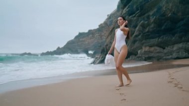 Seksi genç bayan bulutlu deniz kıyısında ıslak kumda yürürken rahatlıyor. Beyaz baştan çıkarıcı mayo giyen çekici, zayıf bir kız. Taşlı kayanın önündeki boş kumsalda poz veriyor. Tasasız kadın güzel okyanusun tadını çıkarıyor..