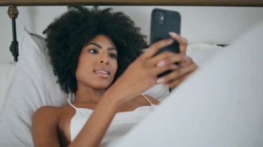 Afrikalı bir kız cep telefonuyla gülümsüyor. Kıvırcık saçlı pozitif kadın elinde telefonla sosyal medyayı okuyor. Afrikalı sağlıklı kadın yumuşak yastığa akıllı telefon kullanarak uzanıyor.