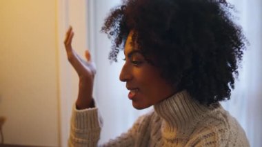 Duygusal bir kadın eve yakın konuşuyor. Kıvırcık saçlı, pozitif kadın akşam apartmanında el hareketleriyle konuşuyor. Afro-Amerikalı mutlu kız beyaz duvarlı oturma odasına video çağrısında bulunan birine gülümsüyor.