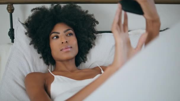 非洲妇女在床上摆出移动自拍的姿势 衣冠楚楚的姑娘把自己的画像放在白色枕头上 微笑的女士在柔软的酒店床垫前拍照 现代女性生活方式概念 — 图库视频影像