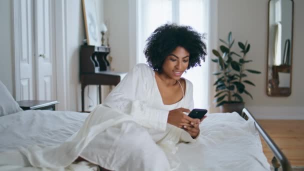 在卧室里笑着给智能手机发短信在酒店内部 卷发女士用手机发短信 非洲裔美国女孩躺在床上享受社交网络设备的通信 — 图库视频影像