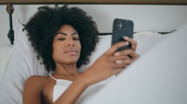 Şehvetli bir kadın yatakta cep telefonunu dinlendiriyor. Yatak odasında fotoğraf çeken neşeli kıvırcık kadın. Afrika kökenli Amerikalı gülümseyen kız uyumadan önce dinleniyor. Modern kadın yaşam tarzı konsepti