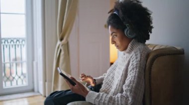 Rahat evdeki odaklanmış kız kaydırma tahtası. Serbest çalışan, tablet seyreden, sakince tek başına çalışan bir kadın. Afro-Amerikan kulaklıklı bayan rahat koltukta oturmuş müzik dinliyor. Ev içi rutin kavram