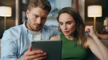 Şaşırmış çift tablet evde zum yapıyor. Genç bir kadın, sıcak kanepe kapanışında kocasına bakıyor. Şok olmuş eşlerin yüzleri, yabancı bir televizyon dizisini izlerken evden yüklü faturalar geliyor.
