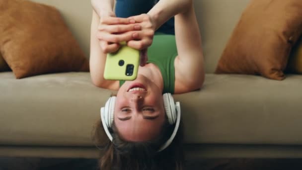 ヘッドフォンの女の子ソファ室内で音楽を聞いて敷設 ホームズームアウトで歌踊りの足を楽しむ幸せな笑顔の女性 週末の家で楽しい女性感ファンキーなテキストメッセージ携帯電話 — ストック動画