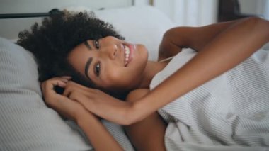 Evde yatan gülümseyen bir portre modeli. Kıvırcık saçlı mutlu kadın sabah yatak odasında uyanmış. Afrika kökenli Amerikalı neşeli kız ortopedik yatakta şekerleme yaptıktan sonra tembel hafta sonunun tadını çıkarıyor.