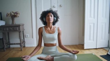 Halıda meditasyon yapan sakin kadın uzaklaşsın. Lotus pozu veren Afro-Amerikan sakin kadın. Genç kıvırcık kadın yoga yapıyor, müzik dinliyor. Kulaklıklarla gevşeyen ince düşünceli kız.