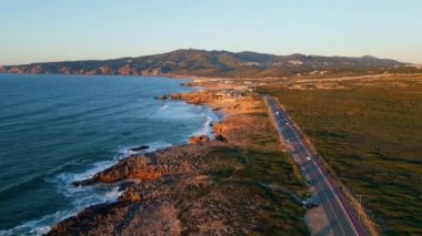 Yazın gün batımında asfalt yolu olan güzel kayalık deniz kıyıları. Taşlı kıyı şeridi boyunca asfalt otobanında uçan hava manzaralı nakliye aracı. Mavi köpüklü okyanus dalgaları sahili yavaş çekimde yıkıyor..