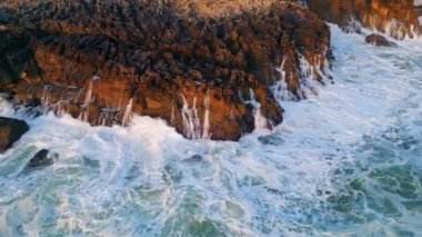 Sert deniz dalgaları taşlı kıyı şeridinde köpürüyor. Fırtınalı deniz dalgaları deniz kıyısı kayaları drone görüntüsünü bozuyor. Güneşli bir günde okyanus manzarası ağır çekimde. Dramatik vahşi deniz manzarası. Turizm konsepti