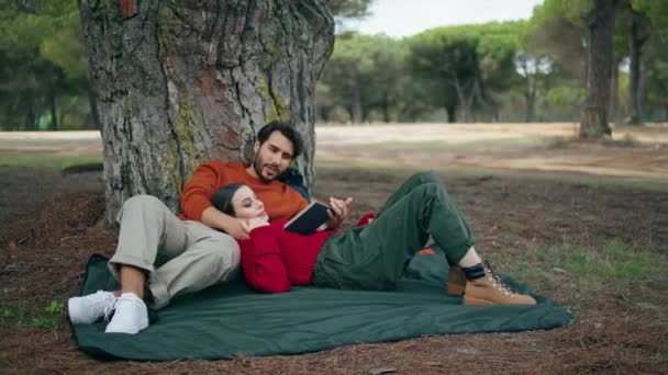 松懈的年轻夫妇躺在宽阔的树下的野餐毛毯上 在秋天的公园里 有魅力的大胡子男人大声地为快乐的女人读书 无忧无虑的家庭对大自然感到寒冷 周末休闲概念 — 图库视频影像