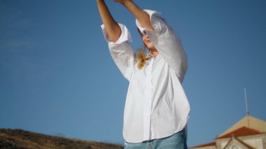 Gözlerini kısarak doğa portresi yapan kız. Sarışın, mutlu kadın kale arkasında tek başına duruyor. Güneş ışınlarını ısıtan kadın model. Yüzü pozitiftir. Yakın plan. Mavi gökyüzünde dinlenen genç bayan