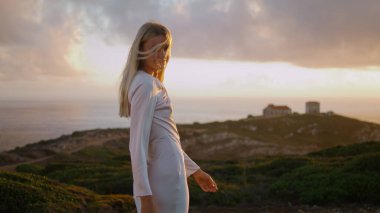 Elbiseli kız akşam deniz kıyısında kameraya bakıyor. Gün batımında şatonun etrafında eli çeken olumlu bir kadın. Sarışın model, yaban rüzgarlı okyanus uçurumlarında dinlenen saçları düzleştirir.