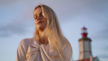 Şehvetli kadın, deniz feneri arka plan portresi çiziyor. Sonbahar akşamları rahat modeller saç modelleri birbirine dokunur. Sarışın kadın kameraya bakıyor manzaralar zum yapıyor. Turizm konsepti