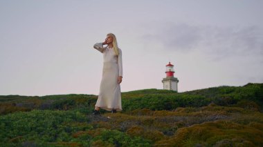 Deniz fenerinin arka planına hayran olan güzel bir kadın. Yeşil doğada poz veren güzel bir kız. İnce kadın model, dışarıda gün batımına benzeyen saçları düzleştiriyor. Sarışın kadın tek başına kameraya bakıyor.