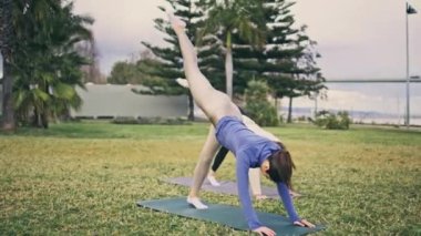 Parkta atletizm takımı antrenmanı. Güçlü bir yogini esneme kuvveti, çimenlerde bir bacağını kaldırıyor. Sportif kadınlar aerobik yoga egzersizleri yapıyor. Partner eğitiminden zevk alıyorlar. Formda kızlar aktif yaşam tarzı 