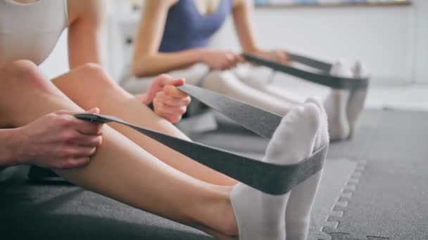 把女人的腿绑在瑜伽垫上 默默无闻的夫妻坐在地毯上 一起享受身体训练 两名灵活的女运动员在白人家庭演播室使用健身弹性带进行锻炼 — 图库视频影像