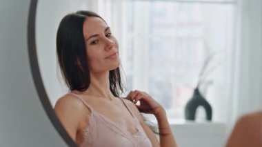 Bakımlı model yumuşak tenli aynaya dokunuyor. Güzelce gülümseyen, saç stili düzelten banyoda yansımaya göz kırpan güzel bir kadın. Evde mutlu kadın saç bakımı. Güzellik konsepti 