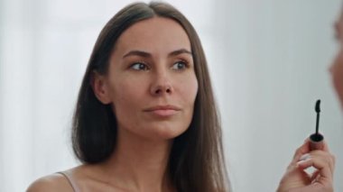 Beyaz mekanda kirpikleri boyamak için kullanılan tatmin edici bir model. Taze bakımlı kadın evde günlük makyaj hazırlıyor. Maskara süren gülümseyen bayan aynaya bakıyor. Güzel kız sabah rutinini evde yapıyor. 