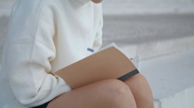 Unknown young woman writing into notebook sitting staircase close up. Girl hands with pen making notes in notepad diary outdoors. Unrecognizable freelancer working sitting on stairs. Urban lifestyle.