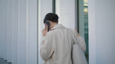 Back view businesswoman calling phone walking near modern office building. Young busy girl manager talking smartphone on go hurrying to work. Elegant confident business lady discussing job by mobile.