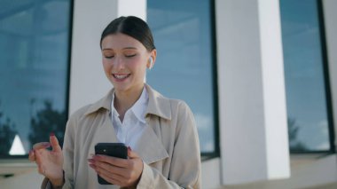 Smiling young woman saying goodbye in phone camera standing city street close up. Elegant attractive girl talking on video call wearing wireless headset outdoors. Happy cheerful lady chatting mobile. 