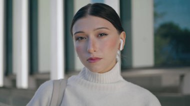 Serious gorgeous businesswoman looking camera confidently standing city street close up. Portrait of attractive young woman wearing modern wireless headphones. Beautiful girl model posing outdoors.