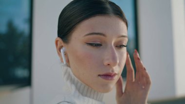 Attractive elegant girl answering call by touching wireless headphones standing city street close up. Portrait of stylish business lady talking phone using modern headset. Young woman listening earbud