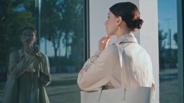Confident young woman looking her reflection in office window taking off wireless headphones outdoors. Attractive business lady standing near city building close up. Businesswoman turning to camera.