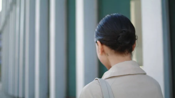 stock image Back view of unknown walking woman straightening hairstyle close up. Unrecognizable young girl going near modern city building wearing stylish coat. Active pretty lady with hair bunch hurrying on work