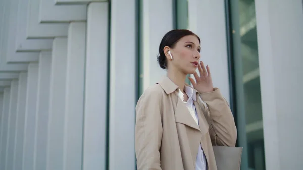 Busy woman speaking wireless earbuds standing near modern city building close up. Confident businesswoman solving work issues remotely talking with modern headset. Elegant girl calling phone outdoors.